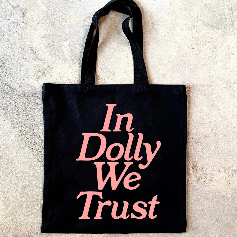 Black tote bag with 'In Dolly We Trust' text in pink on a light gray background
